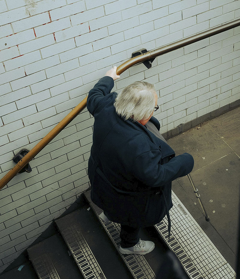 Vista desde arriba de una señora mayor subiendo unas escaleras ayudándose de la barandilla.